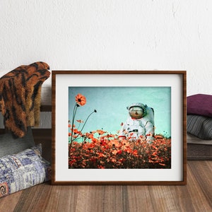 K&ouml;nnte beinhalten: Ein gerahmtes Foto eines Astronauten, der in einem Feld mit orangefarbenen Blumen steht. Der Astronaut tr&auml;gt einen wei&szlig;en Raumanzug mit einem blauen Helm. Der Hintergrund ist ein hellblauer Himmel.