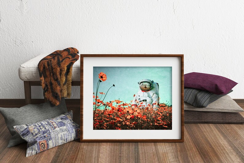 K&ouml;nnte beinhalten: Ein gerahmtes Foto eines Astronauten, der in einem Feld mit orangefarbenen Blumen steht. Der Astronaut tr&auml;gt einen wei&szlig;en Raumanzug mit einem blauen Helm. Der Hintergrund ist ein hellblauer Himmel.