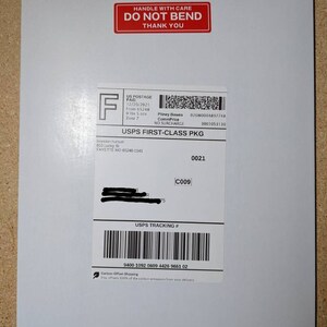 K&ouml;nnte beinhalten: Ein wei&szlig;er Umschlag mit einem roten "Vorsichtig behandeln, Nicht knicken, Danke"-Etikett. Der Umschlag hat ein USPS First-Class Package-Etikett mit einer Sendungsverfolgungsnummer, "9400 1092 0609 4426 9661 02", und ein Logo f&uuml;r CO2-kompensierten Versand.