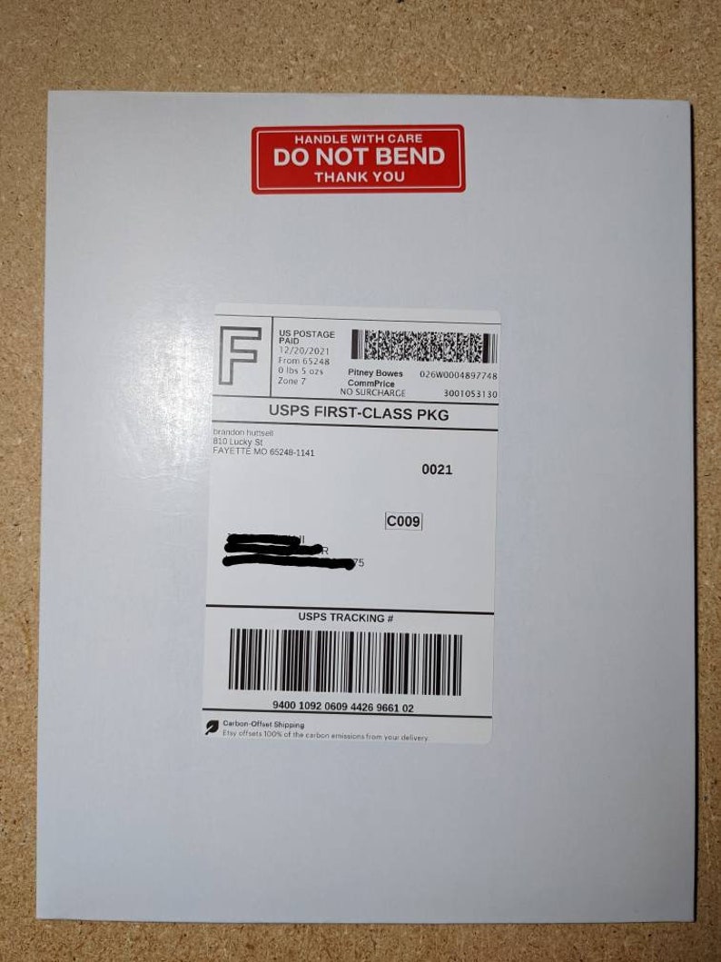 K&ouml;nnte beinhalten: Ein wei&szlig;er Umschlag mit einem roten "Vorsichtig behandeln, Nicht knicken, Danke"-Etikett. Der Umschlag hat ein USPS First-Class Package-Etikett mit einer Sendungsverfolgungsnummer, "9400 1092 0609 4426 9661 02", und ein Logo f&uuml;r CO2-kompensierten Versand.