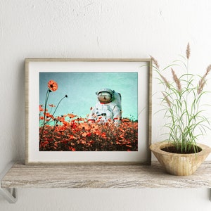 K&ouml;nnte beinhalten: Ein gerahmter Druck eines Astronauten, der in einem Feld mit orangefarbenen Blumen vor blauem Himmel sitzt.