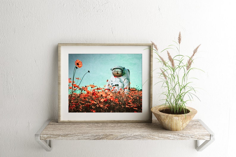 K&ouml;nnte beinhalten: Ein gerahmter Druck eines Astronauten, der in einem Feld mit orangefarbenen Blumen vor blauem Himmel sitzt.