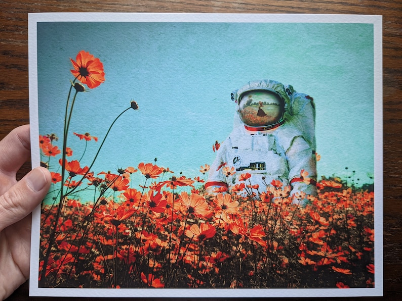 K&ouml;nnte beinhalten: Ein Astronaut in einem wei&szlig;en Raumanzug steht in einem Feld mit orangefarbenen Kosmosblumen. Das Visier des Astronauten spiegelt eine Szene einer Person wider, die in einem Feld geht.