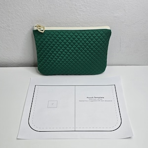 May include: A quilted emerald green pouch with a gold zipper pull and a white trim. A paper template labeled "Pouch Template" is below the pouch. The template includes a 1-inch square and seam allowance instructions.