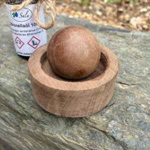 May include: A wooden aromatherapy diffuser with a round ball and a small bowl. A small bottle of citronella oil is in the background. The bottle is wrapped with twine and has a label with text.