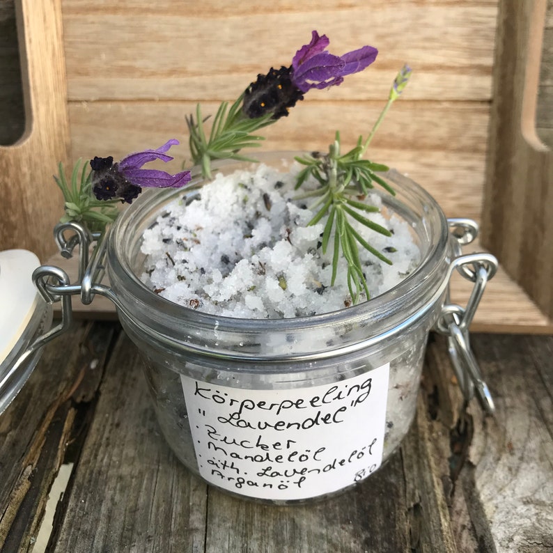 May include: A glass jar filled with a white sugar scrub with lavender and rosemary. The jar has a handwritten label that reads "K&ouml;rperpeeling "Lavendel" : Zucker : Mandel&ouml;l : &auml;th. Lavendel&ouml;l : Argan&ouml;l"