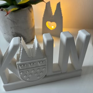 May include: A white concrete sculpture spelling out "KOLN" with a detailed depiction of the Cologne Cathedral. A small, illuminated cathedral-shaped candle holder with a heart cutout is in the background.