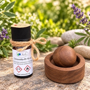 Op de afbeelding: Een flesje van 10 ml citronella-olie met een zwarte dop, omwikkeld met touw, voorzien van een wit etiket met tekst en gevarensymbolen. Daarnaast ligt een gladde, ronde houten bol in een ronde houten voet op een gestructureerde ondergrond.