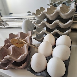 Op de afbeelding: Een stapel grijze en roze eierdozen. Zes witte eieren liggen in een donkergrijze doos. Een klein label met de tekst "Handmade with love" is bevestigd aan de roze dozen.