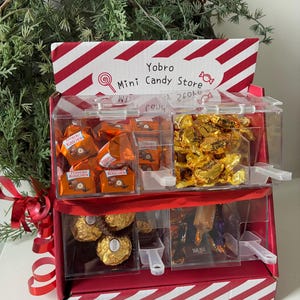 Puede incluir: Un expositor de mini tienda de caramelos a rayas rojas y blancas con compartimentos transparentes llenos de caramelos envueltos. La parte superior de la pantalla tiene el texto "Yobro Mini Candy Store". Un lazo rojo y vegetación en el fondo.
