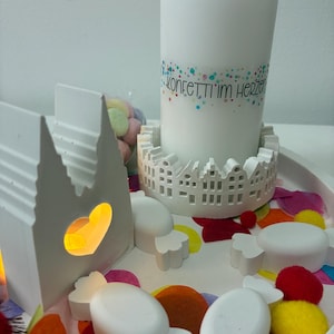 Può includere: Una candela bianca con la scritta "Konfetti im Herzen" è posta in un supporto decorativo. La scena comprende decorazioni bianche a forma di caramella, una luce a forma di cuore e coriandoli colorati.