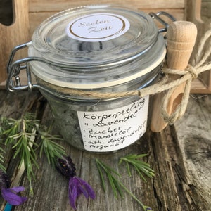 May include: A glass jar with a metal lid filled with a lavender body scrub. The jar has a label that reads "K&ouml;rperpeeling "Lavendel"  Zucker, Mandel&ouml;l, &auml;th. Lavendel&ouml;l, Argan&ouml;l".