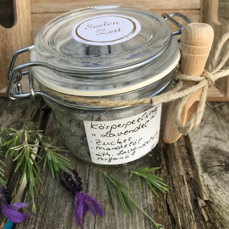 May include: A glass jar with a metal lid filled with a lavender body scrub. The jar has a label that reads "K&ouml;rperpeeling "Lavendel"  Zucker, Mandel&ouml;l, &auml;th. Lavendel&ouml;l, Argan&ouml;l".