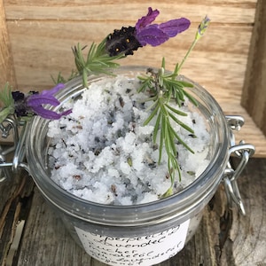 May include: A glass jar filled with white sugar scrub, lavender buds, and rosemary sprigs. The jar has a metal clasp and a label that reads "Perfecting Lavender Sugar Scrub".