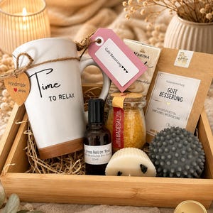 Könnte beinhalten: Ein Geschenkset in einer Holzschale mit einer weißen Tasse mit der Aufschrift Time TO RELAX, einem 20ml Rosen-Roll-on, einem Glas gelbem Badesalz, einer weißen Kerze, einem grauen Noppenball und einer Teepackung mit der Aufschrift GUTE BESSERUNG.