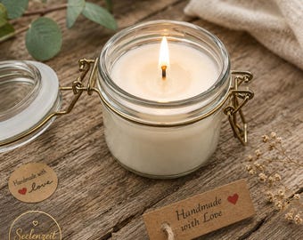 May include: A white candle in a clear glass jar with a metal wire bail closure. The candle is lit with a visible flame. A brown paper tag attached with twine reads Handmade with Love and features a small red heart icon printed on the surface.