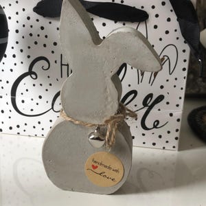 Può includere: Una statuina di coniglio in cemento grigio con lunghe orecchie e un corpo rotondo. Il coniglio è legato con spago e una piccola campana d'argento. È attaccato un adesivo rotondo con le parole "handmade with love". Lo sfondo include un sacchetto regalo a pois.