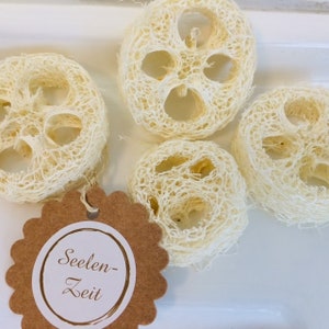 May include: Four natural loofah sponges in a white bowl. The loofahs are round and have a woven texture. A brown tag with white text reads "Seelen-Zeit".