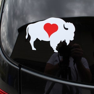 May include: A white silhouette of a buffalo with a red heart in the center, on a black car window.