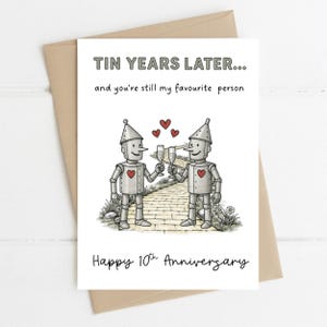 Könnte beinhalten: Eine Grußkarte zum 10. Jahrestag. Die Karte zeigt zwei Zinnroboter, die mit Champagnergläsern anstoßen. Der Text lautet: "Tin Years Later... and you're still my favourite person" und "Happy 10th Anniversary".