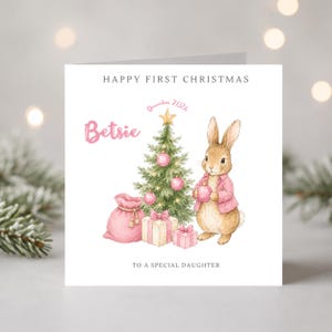 Könnte beinhalten: Eine quadratische Grußkarte mit einer Aquarellillustration eines braunen Kaninchens in einer rosa Jacke neben einem geschmückten Weihnachtsbaum, Geschenken und einem rosa Sack. Der Text lautet HAPPY FIRST CHRISTMAS, Betsie, December 2026, TO A SPECIAL DAUGHTER.