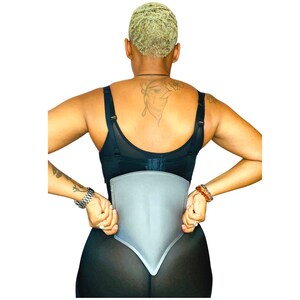 May include: A black and gray waist trainer with a smooth, contoured design. The trainer is worn by a person with short, blonde hair.