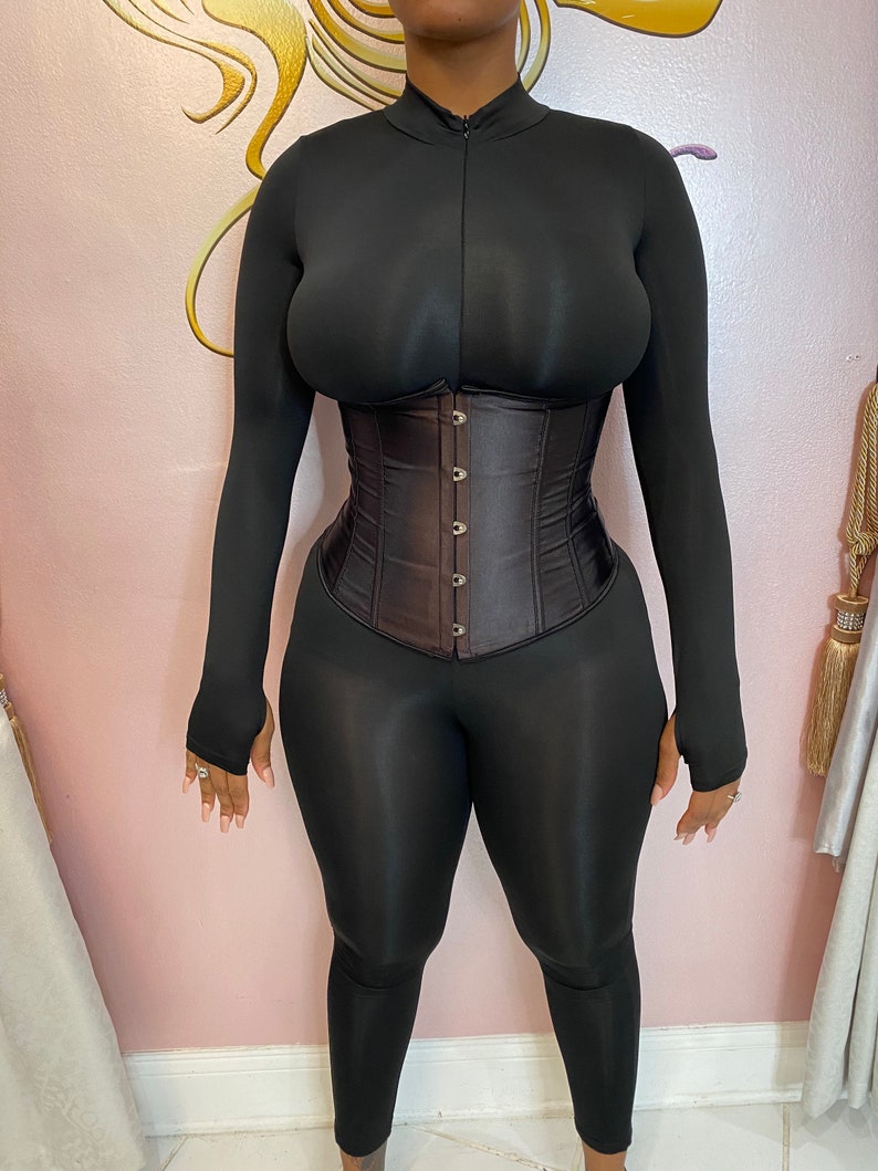 K&ouml;nnte beinhalten: Ein schwarzer Langarm-Body mit einem braunen Satin-Korsett. Der Body hat einen Stehkragen und einen Rei&szlig;verschluss. Das Korsett hat einen Busk-Verschluss und Metall&ouml;sen.