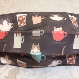 以下が含まれることがあります： コーヒーカップに入った猫が繰り返し描かれた、濃い茶色の布製のマスク。マスクには「カプチーノ」「エスプレッソ」「モカ ラテ」「コールドブリュー」「コーヒー オブ ザ デイ」の文字が書かれている。