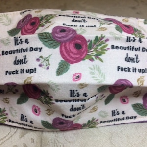 Pode incluir: Uma máscara facial de tecido branco com um padrão floral e o texto "It's a Beautiful Day don't Fuck it up!" repetido em toda a superfície.