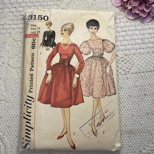 May include: A vintage Simplicity sewing pattern packet with illustrations of women's dresses. The packet is labelled "3150" and includes sizing information. The dresses are in shades of red and pink, with a black dress on the side. A pink flower is in the background.