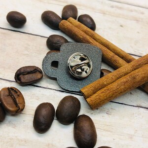 May include: A gray metal coffee cup pin with a silver pin back. The pin is surrounded by cinnamon sticks and coffee beans.