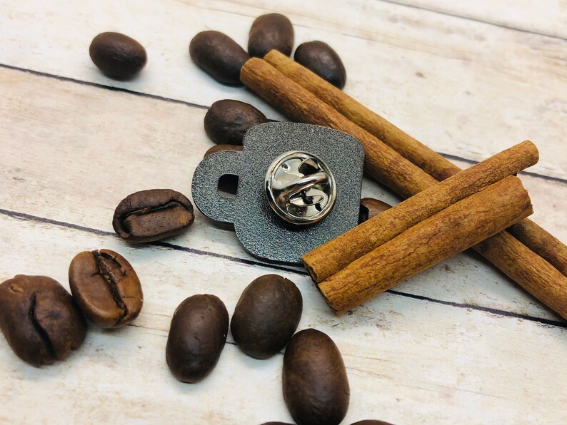 May include: A gray metal coffee cup pin with a silver pin back. The pin is surrounded by cinnamon sticks and coffee beans.