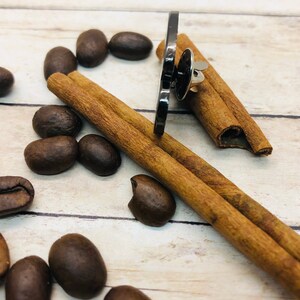 May include: A black metal pin with a silver pin back, surrounded by coffee beans and cinnamon sticks on a light wood surface.