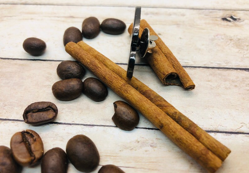 May include: A black metal pin with a silver pin back, surrounded by coffee beans and cinnamon sticks on a light wood surface.