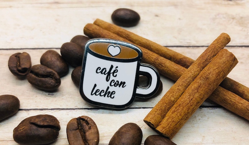 May include: A silver enamel pin shaped like a coffee mug with the text "caf&eacute; con leche" and a small white heart on top. The pin is surrounded by cinnamon sticks and coffee beans.