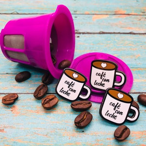 May include: Three enamel pins shaped like coffee mugs with the text "caf&eacute; con leche" in black and white. The pins are on a blue and white wooden surface with coffee beans scattered around them.