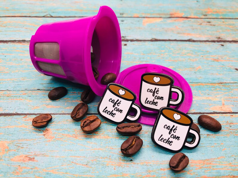 May include: Three enamel pins shaped like coffee mugs with the text "caf&eacute; con leche" in black and white. The pins are on a blue and white wooden surface with coffee beans scattered around them.