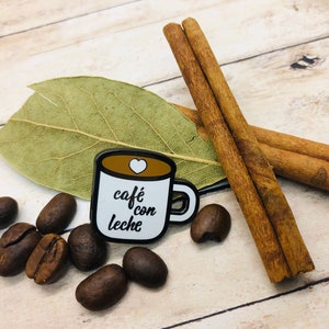 May include: A white enamel pin shaped like a coffee mug with the text "caf&eacute; con leche" and a small heart on the side. The pin is surrounded by coffee beans, cinnamon sticks, and a bay leaf on a wooden surface.