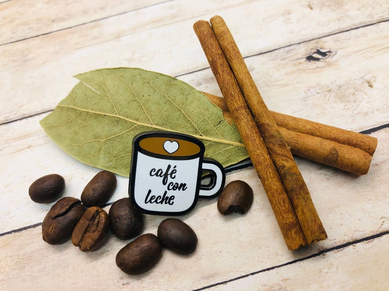 May include: A white enamel pin shaped like a coffee mug with the text "caf&eacute; con leche" and a small heart on the side. The pin is surrounded by coffee beans, cinnamon sticks, and a bay leaf on a wooden surface.