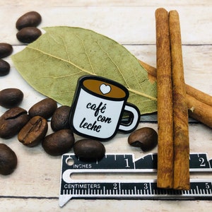 May include: A white enamel pin shaped like a coffee mug with the text "caf&eacute; con leche" in black cursive writing and a small red heart on the top. The pin is surrounded by coffee beans and cinnamon sticks.