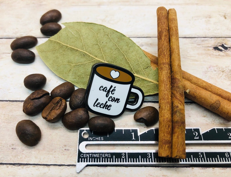 May include: A white enamel pin shaped like a coffee mug with the text "caf&eacute; con leche" in black cursive writing and a small red heart on the top. The pin is surrounded by coffee beans and cinnamon sticks.