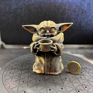 K&ouml;nnte beinhalten: Bronze farbene Figur einer Figur aus einer beliebten Science-Fiction-Serie, die eine kleine Tasse h&auml;lt. Die Figur hat gro&szlig;e Ohren und tr&auml;gt eine Robe. Eine M&uuml;nze befindet sich neben der Figur.