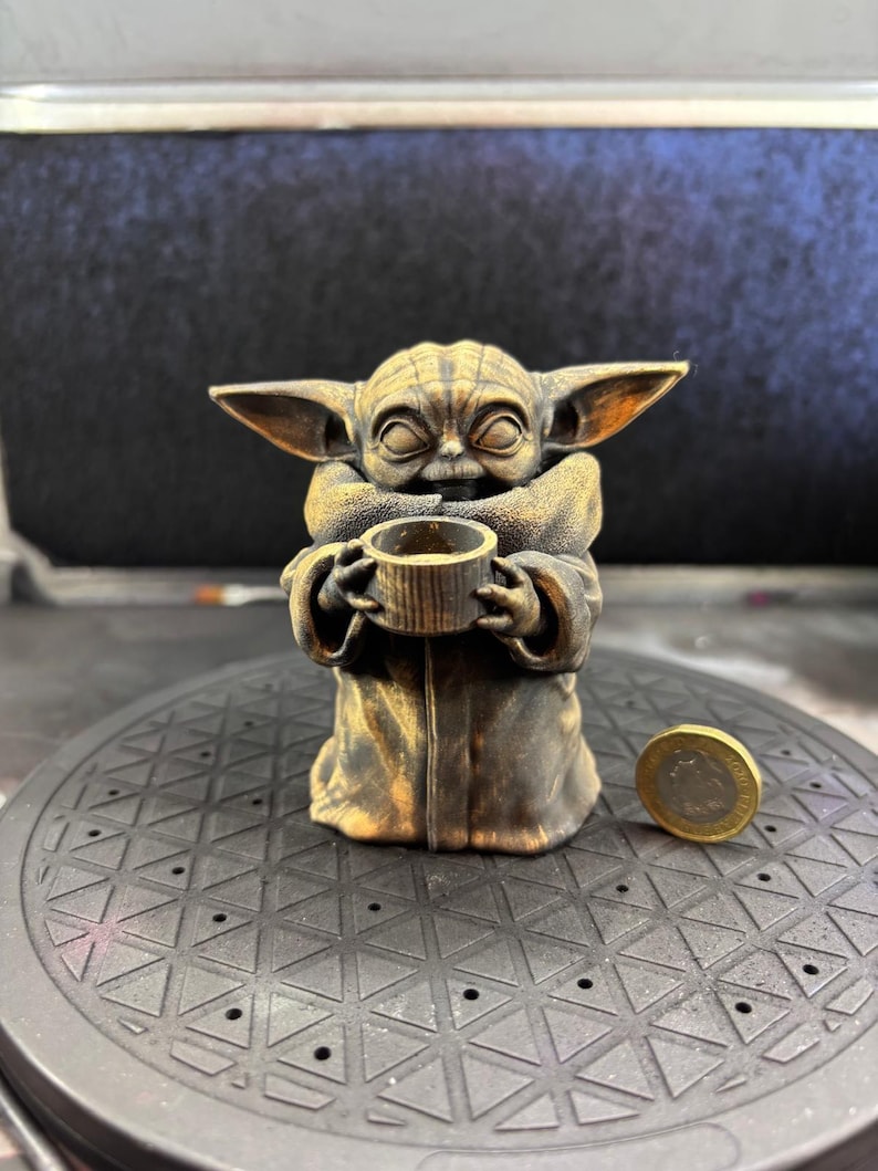 K&ouml;nnte beinhalten: Bronze farbene Figur einer Figur aus einer beliebten Science-Fiction-Serie, die eine kleine Tasse h&auml;lt. Die Figur hat gro&szlig;e Ohren und tr&auml;gt eine Robe. Eine M&uuml;nze befindet sich neben der Figur.