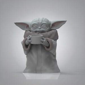 K&ouml;nnte beinhalten: Graue Figur einer Figur aus einem beliebten Science-Fiction-Franchise, die eine kleine Tasse h&auml;lt. Die Figur hat gro&szlig;e Ohren, eine zerknitterte Stirn und tr&auml;gt eine Robe mit einem fellartigen Kragen. Die Figur steht auf einer wei&szlig;en Oberfl&auml;che.