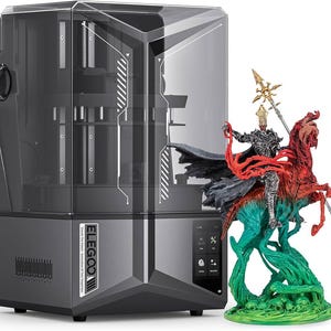 Op de afbeelding: Een zwarte 3D-printer met een transparante kunststof behuizing. De printer staat op een wit oppervlak. Voor de printer staat een 3D-geprint beeldje van een ridder die een draak berijdt. Het beeldje is geschilderd in tinten rood, groen en goud.