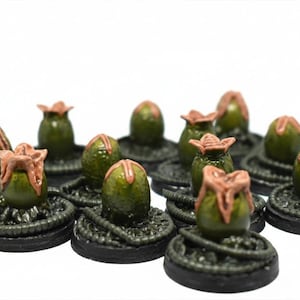 May include: A group of miniature alien eggs. Each egg has a dark green, oval body and a light brown, flower-like top. They are set on a dark, textured base, creating a sci-fi or fantasy theme.