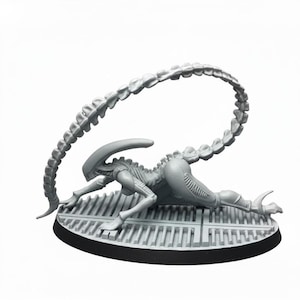 May include: A grey Xenomorph figure, posed on a textured platform with a black base. The creature has a long, segmented tail arching over its back. The figure is detailed with a smooth, sleek body.