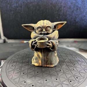 K&ouml;nnte beinhalten: Eine bronzefarbene Figur eines Charakters aus einem beliebten Science-Fiction-Franchise. Die Figur h&auml;lt eine kleine Tasse. Die Figur hat gro&szlig;e Ohren und tr&auml;gt eine Robe. Die Figur sitzt auf einer schwarzen, gemusterten Oberfl&auml;che.