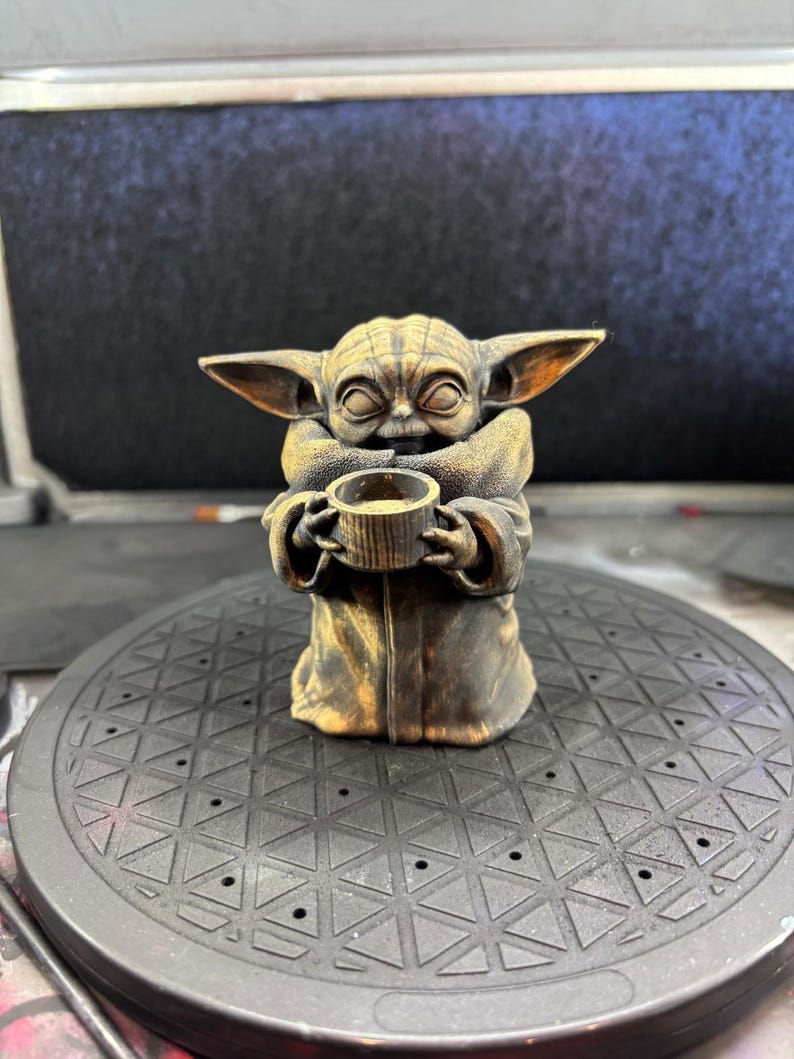 K&ouml;nnte beinhalten: Eine bronzefarbene Figur eines Charakters aus einem beliebten Science-Fiction-Franchise. Die Figur h&auml;lt eine kleine Tasse. Die Figur hat gro&szlig;e Ohren und tr&auml;gt eine Robe. Die Figur sitzt auf einer schwarzen, gemusterten Oberfl&auml;che.