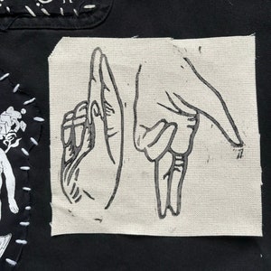 May include: A black and white illustration of two hands making a gesture on a white canvas patch. The patch is sewn onto a black fabric background.